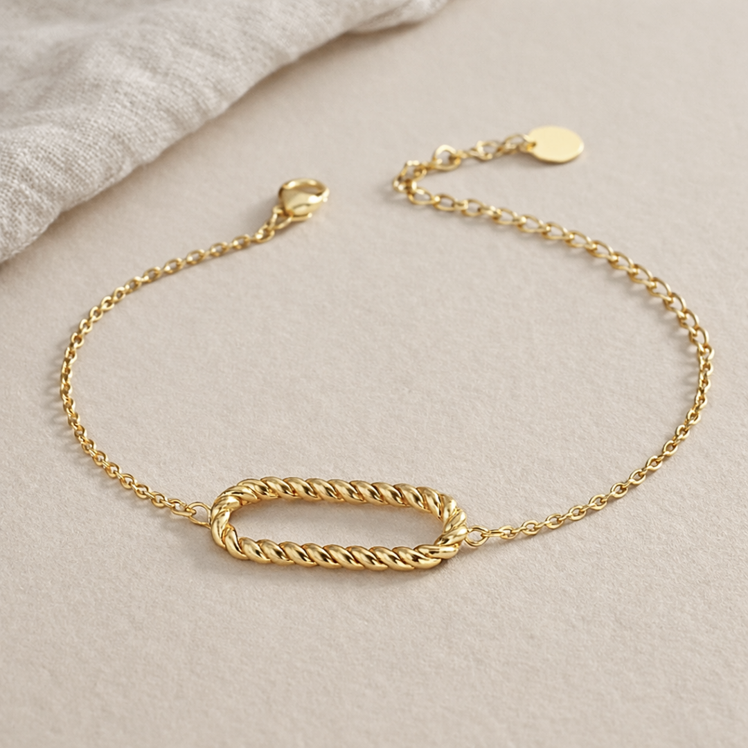 Bracelet fin en métal doré composé d’une chaîne délicate avec au centre un motif ovale torsadé. Fermoir mousqueton avec chaînette d’extension, posé sur un fond blanc texturé.